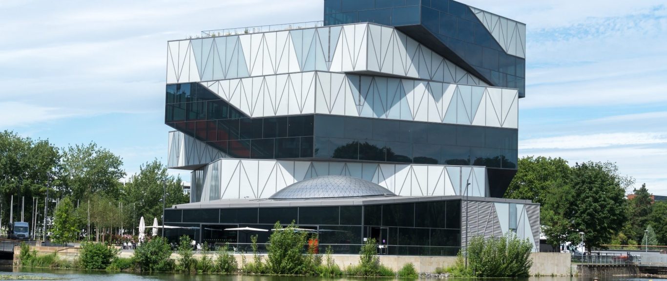 Modernes, geometrisches Gebäude mit Glasfassade, umgeben von Grün und Wasser.