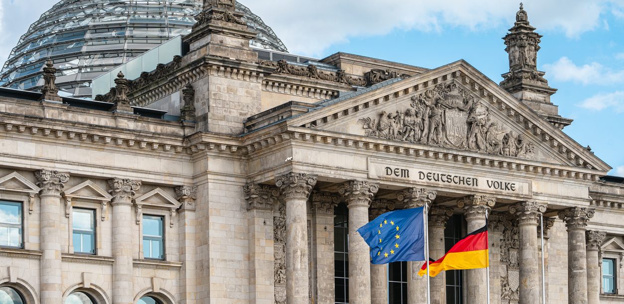 Reichstaggebäude in Berlin mit EU- und Deutscher Flagge vor einer wolkigen Skyline.